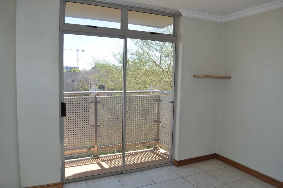 A bright, empty bedroom featuring tiled floors, a floating shelf, and a sliding glass door leading to a private balcony with a metal balustrade.