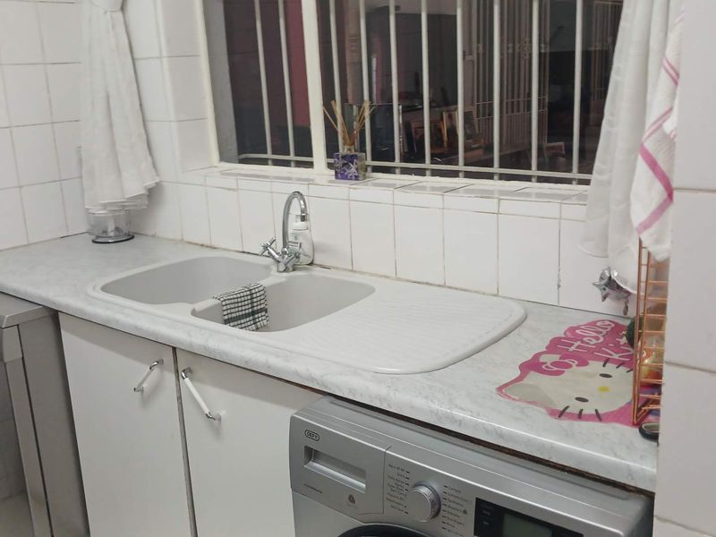 A functional kitchen area featuring a double basin sink, tiled splashback, and laminate countertops with built-in cabinetry and security bars on the window.
