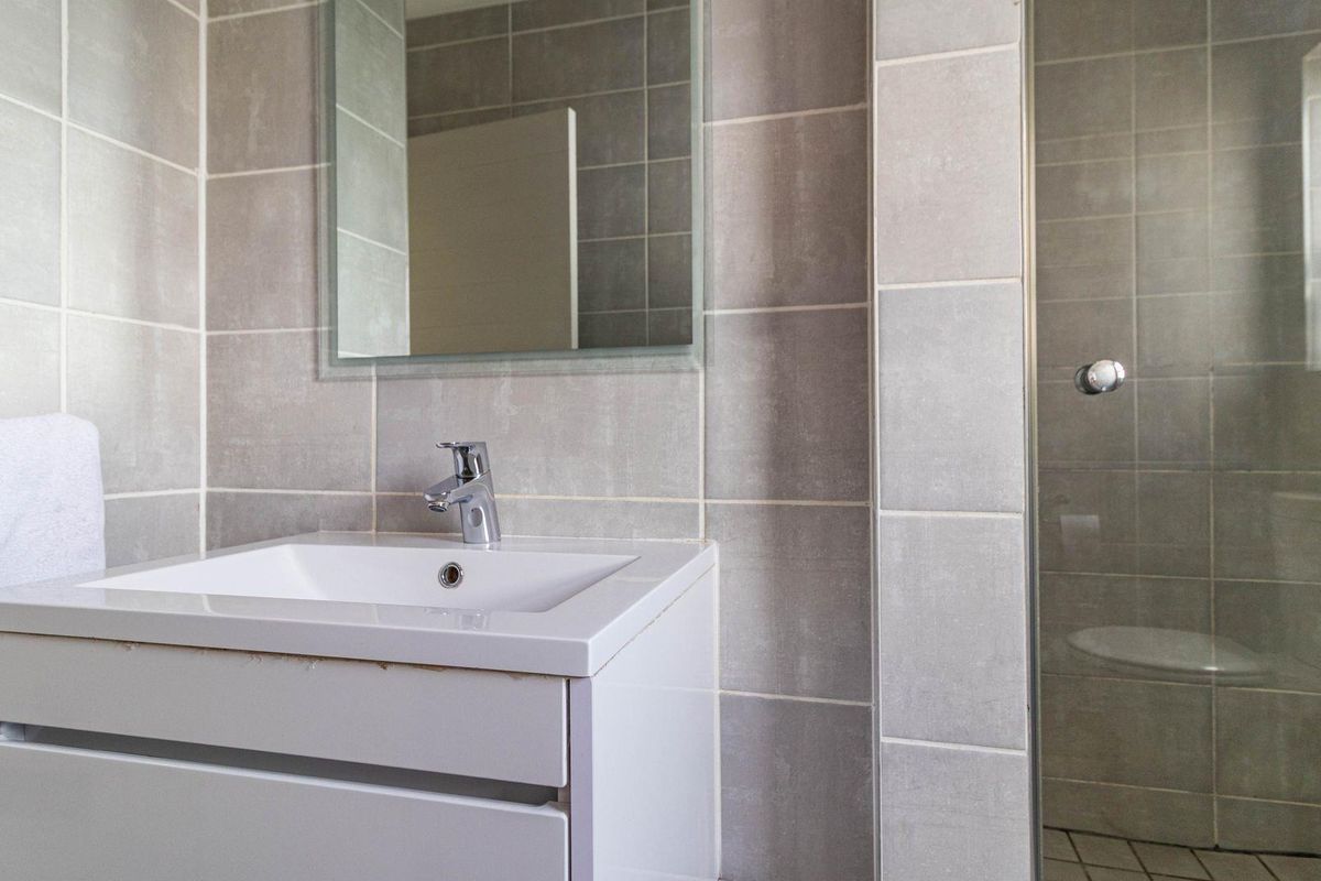 Modern bathroom featuring a sleek white vanity with integrated basin, wall-mounted mirror, and a glass-enclosed shower with neutral grey wall tiling.