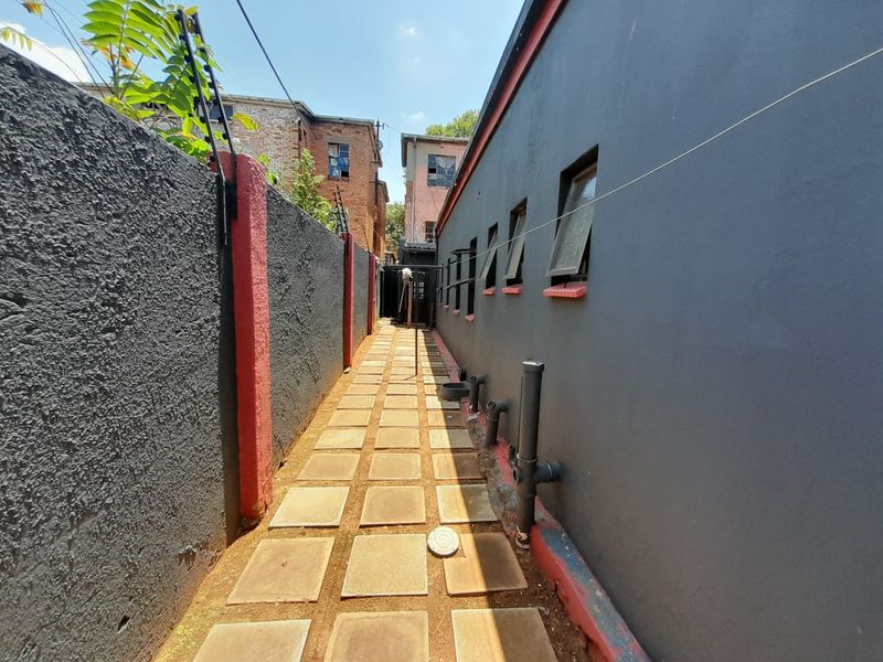 A narrow side passage featuring a paved walkway, boundary wall with electric fencing, and exterior wall access with exposed plumbing.