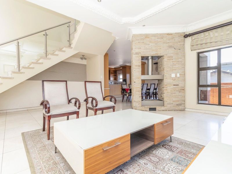 Spacious open-plan living area featuring a central stone-clad fireplace, elegant staircase with glass balustrade, and bright tiled flooring throughout.