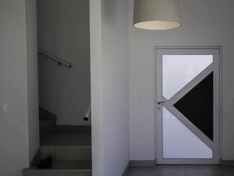 A minimalist entrance hall featuring tiled flooring, a modern geometric glass door, and a staircase with a wall-mounted handrail.