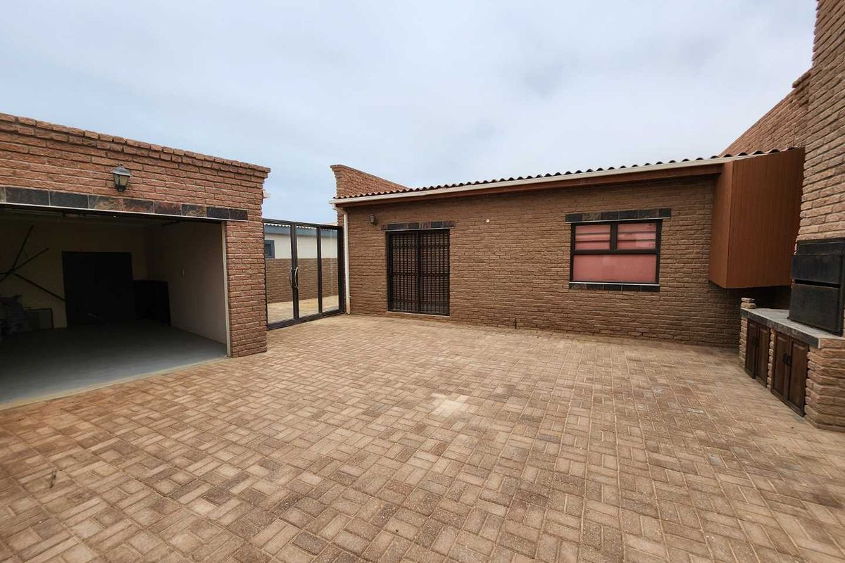 A paved rear courtyard featuring a built-in braai area, brick facade, and access to a garage and main dwelling.