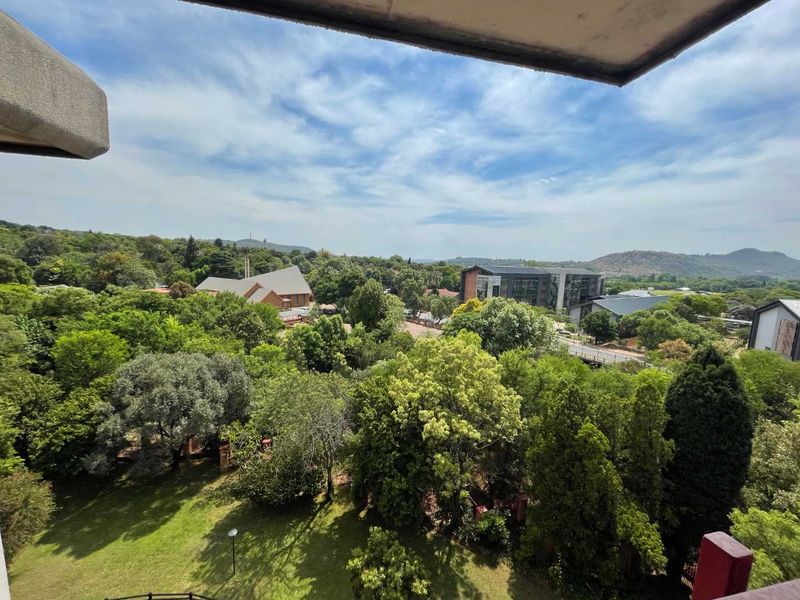 An elevated outdoor view overlooking a lush green landscape with mature trees, nearby buildings, and a clear blue sky.