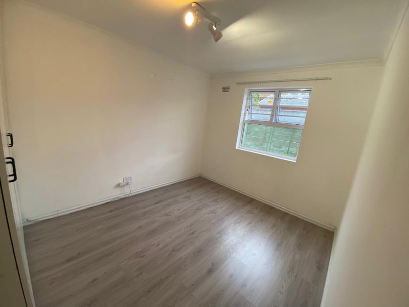 A clean, empty bedroom featuring modern laminate flooring, a window with security bars, and a ceiling-mounted spotlight fixture.