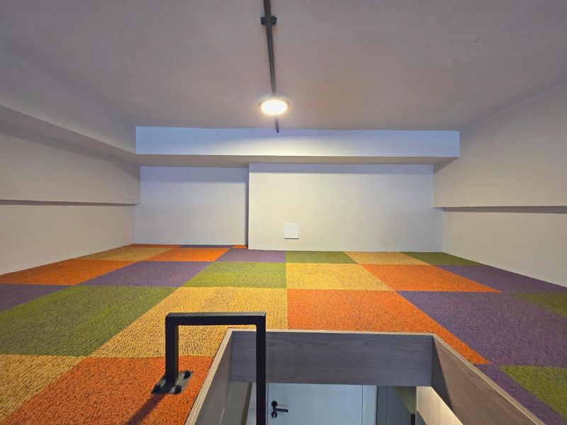 A bright mezzanine level featuring colorful carpet tiles, a minimalist ceiling light, and a secure staircase railing, offering a versatile open-plan loft space.
