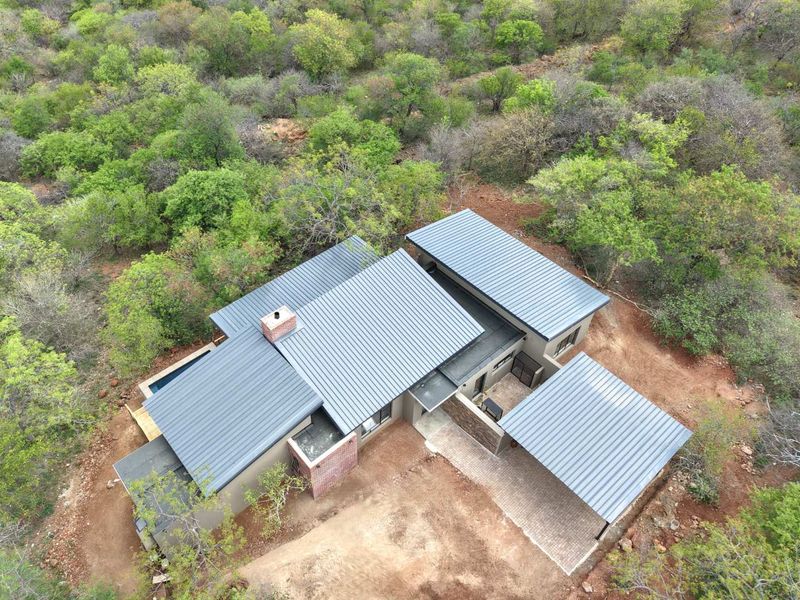Aerial view of a modern house with a metal roof, brick chimney, paved driveway, carport, and swimming pool, set within a natural bushveld environment.