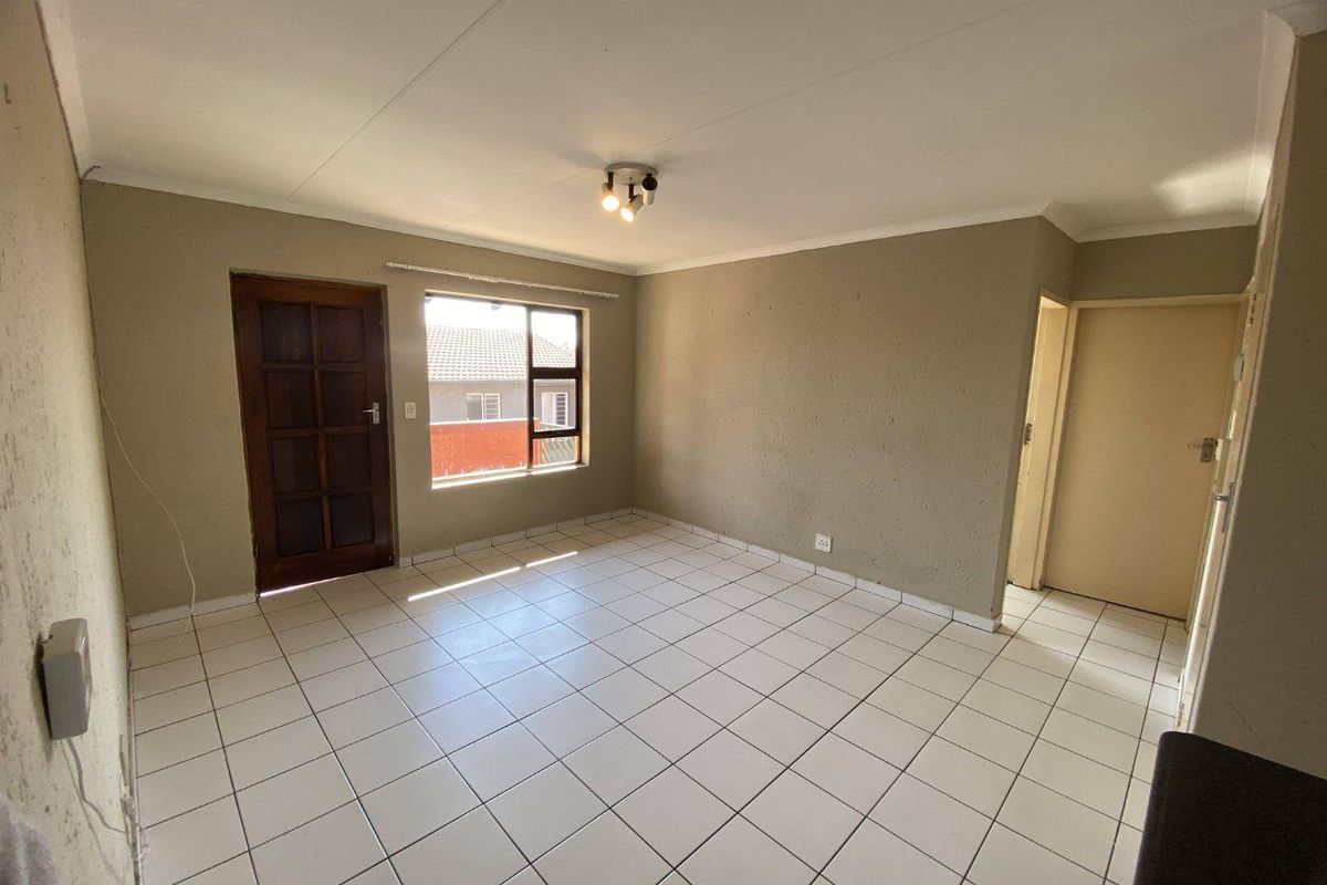 An empty living room featuring white tiled flooring, neutral wall tones, and a timber entrance door with natural light from the window.
