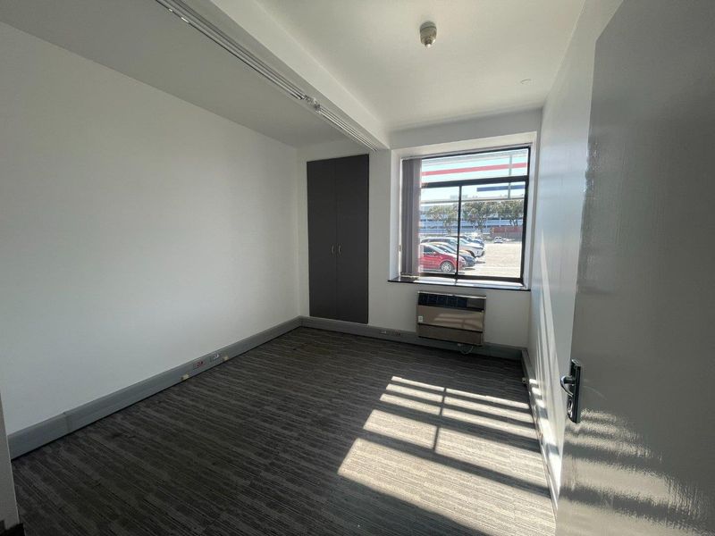 A clean, empty office space featuring carpeted flooring, a built-in cupboard, and a window-mounted air conditioning unit with natural light.