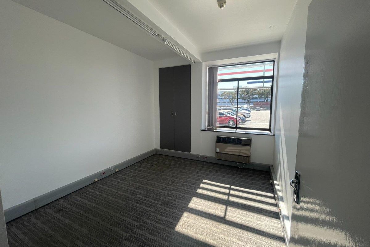 A clean, empty office space featuring carpeted flooring, a built-in cupboard, and a window-mounted air conditioning unit with natural light.