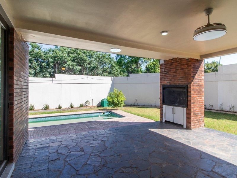 Covered patio featuring stone paving, a built-in braai, and a ceiling fan, overlooking a private swimming pool and garden area.