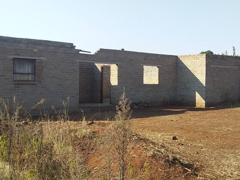 An unfinished residential structure featuring exposed brick masonry walls and open window and door frames, situated on a vacant plot of land.