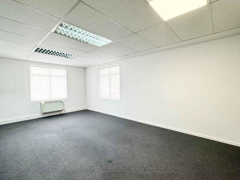 A clean, vacant office space featuring neutral walls, carpeted flooring, suspended ceiling with recessed lighting, and a built-in window air conditioning unit.