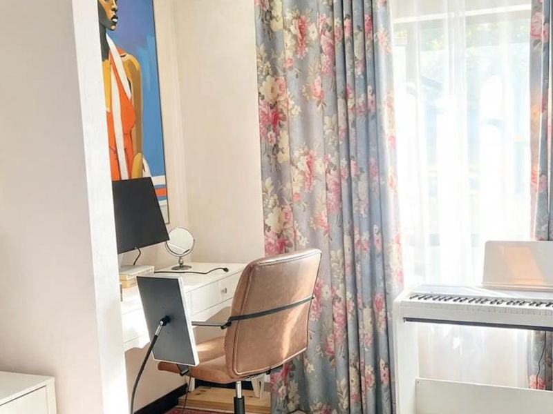 A bright bedroom featuring tiled flooring and a large window dressed with floral curtains, providing a functional space for a home office setup.