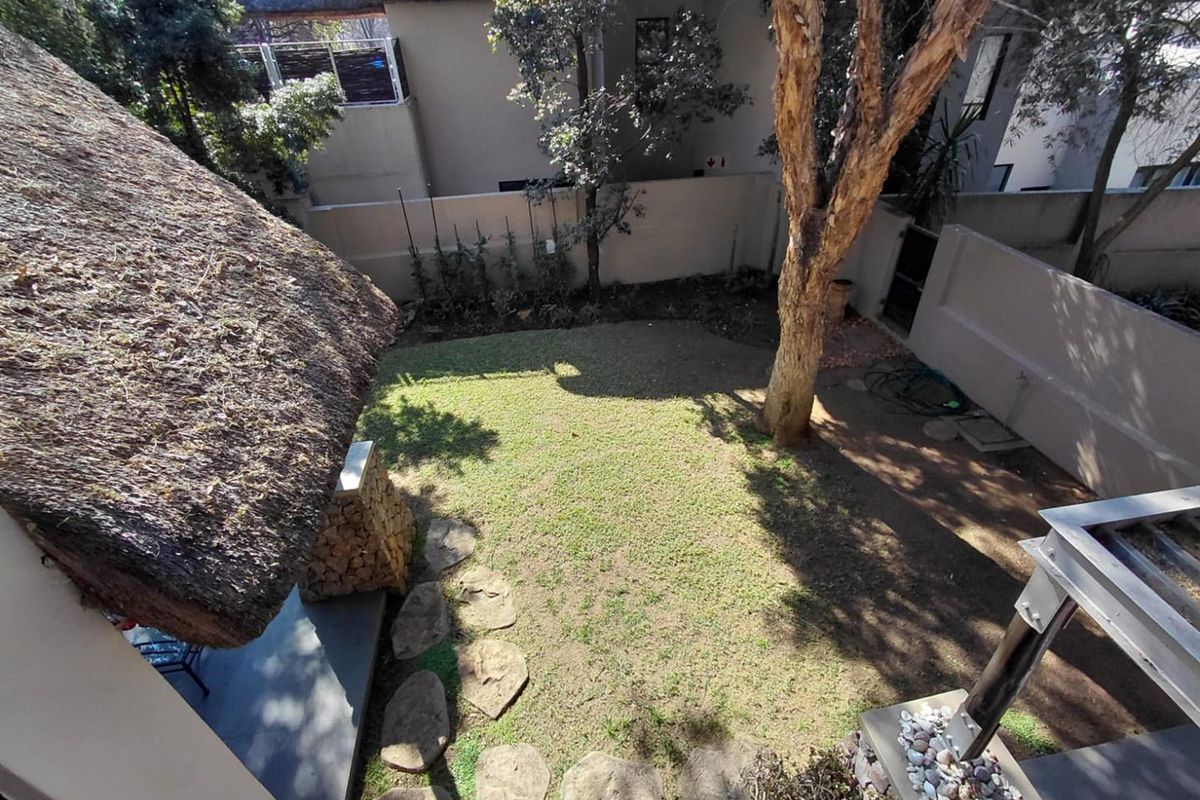 An elevated view of a private rear garden featuring a thatched roof structure, a well-maintained lawn, and established trees enclosed by boundary walls.