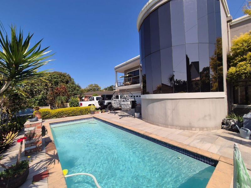 Outdoor pool area featuring a swimming pool with tiled surround, adjacent to a modern home with a distinctive curved glass facade and balcony.