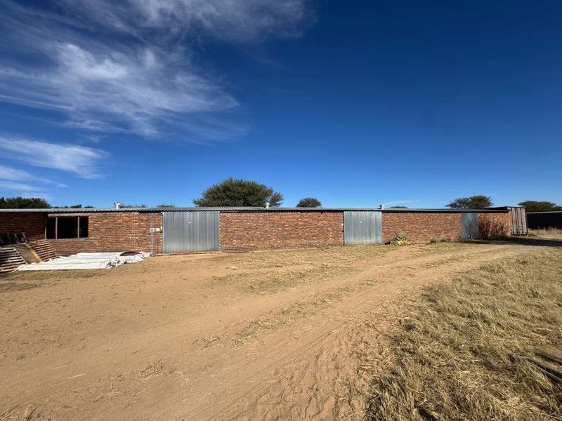 A long, single-story brick outbuilding or storage structure featuring multiple metal roller doors and a corrugated roof, situated on a large, unpaved plot.