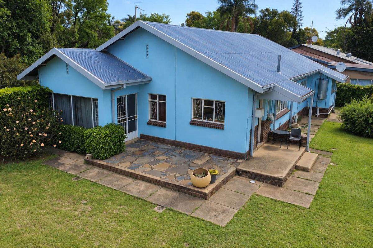 Rear view of the house featuring a blue exterior, corrugated metal roof, and a stone-paved patio area set within a spacious garden.