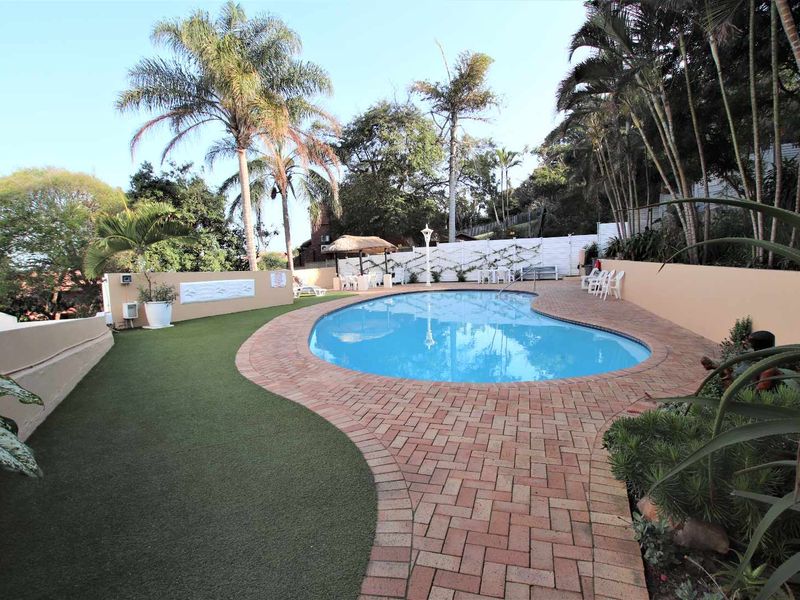 A well-maintained outdoor pool area featuring a curved swimming pool, brick paved surround, artificial lawn, and established perimeter landscaping.