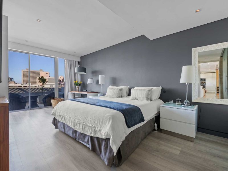 A modern bedroom featuring light laminate flooring, a dark accent wall, and large sliding glass doors providing access to a private balcony with city views.