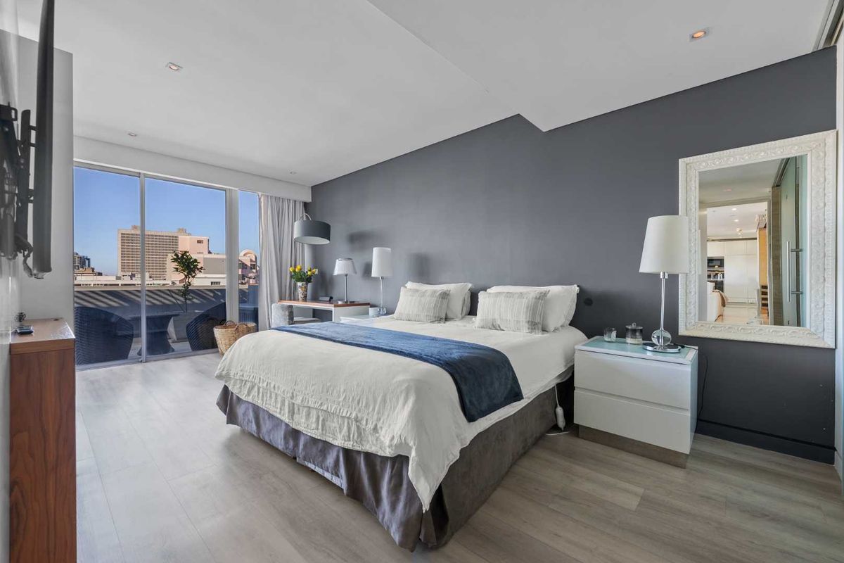 A modern bedroom featuring light laminate flooring, a dark accent wall, and large sliding glass doors providing access to a private balcony with city views.