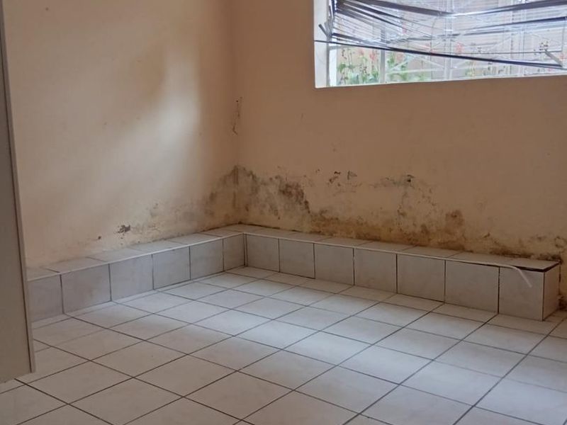 An empty bedroom featuring tiled flooring, an exposed ceiling beam, and a window with security bars, currently showing significant signs of wall dampness.