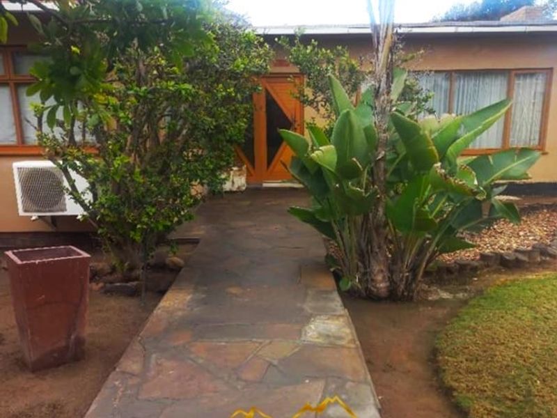 A rear exterior view of the property featuring a stone paved walkway leading to a wooden entrance door and established garden landscaping.