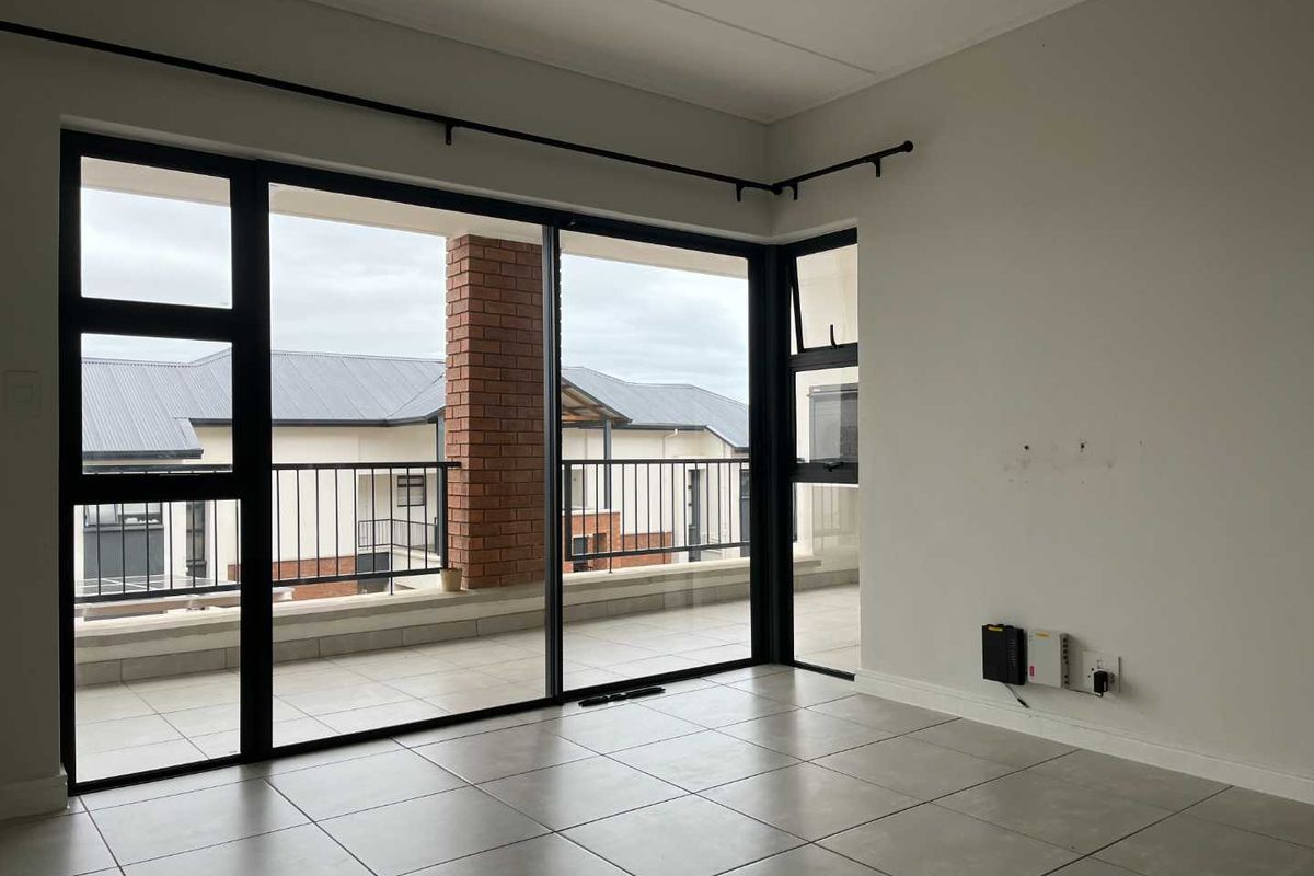 A bright, empty living area featuring large black-framed sliding glass doors that open onto a tiled balcony with metal railings.