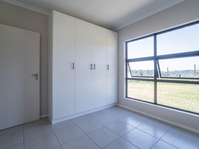 A bright, empty bedroom featuring modern built-in cupboards, neutral tiled flooring, and large aluminium-framed windows overlooking the garden.