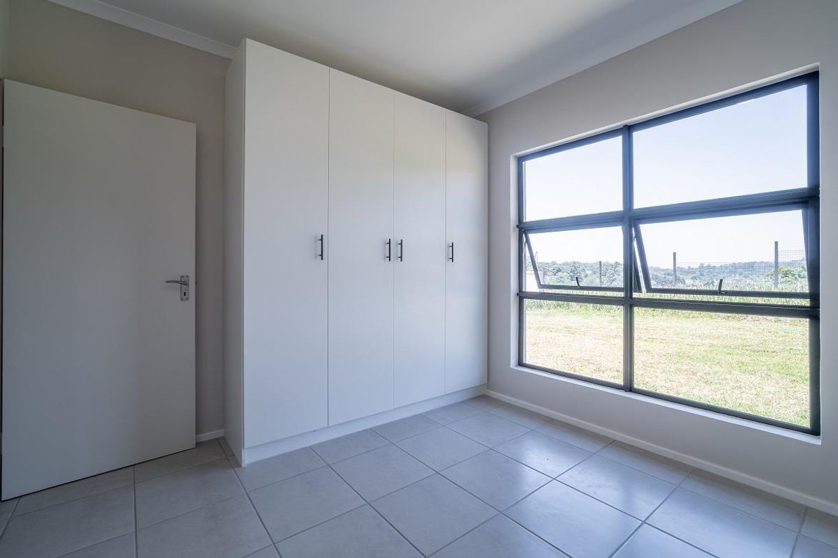 A bright, empty bedroom featuring modern built-in cupboards, neutral tiled flooring, and large aluminium-framed windows overlooking the garden.