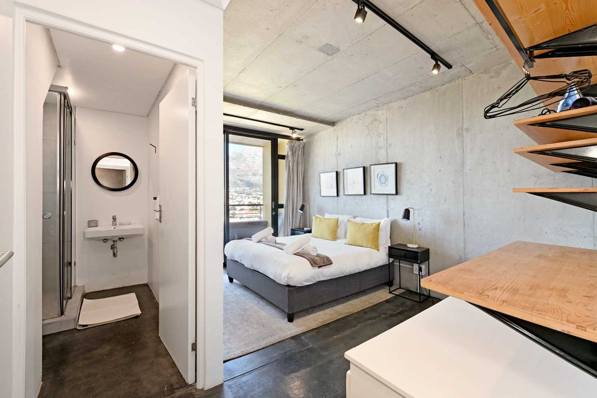 Modern bedroom featuring industrial-style exposed concrete finishes, track lighting, polished concrete floors, and an integrated en-suite bathroom with a glass shower.