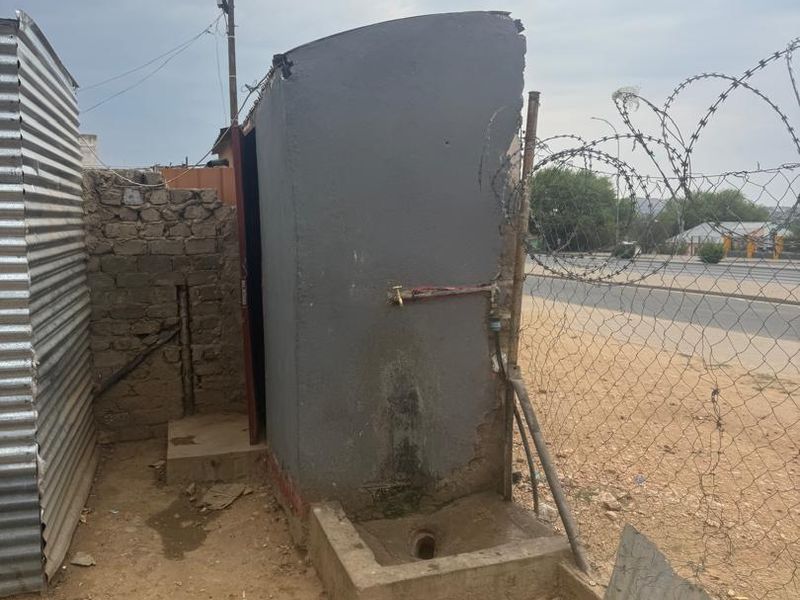 An outdoor utility area featuring a concrete drainage basin, external water tap, and basic masonry and corrugated metal boundary structures.