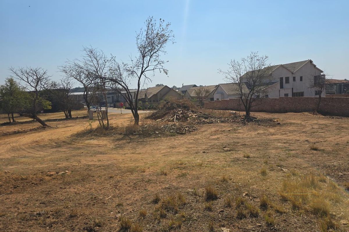 A vacant residential plot featuring natural vegetation, a boundary wall, and surrounding neighborhood housing.