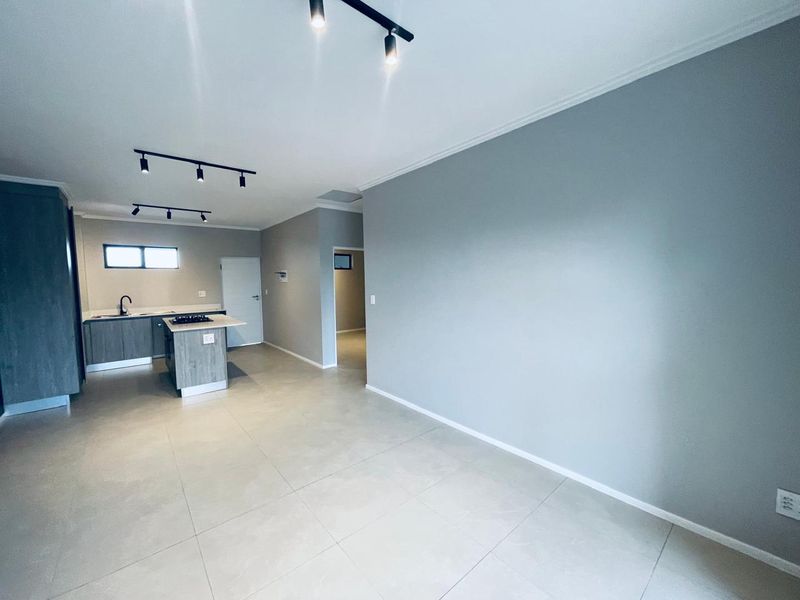 A modern open-plan living and kitchen area featuring neutral tiled flooring, contemporary grey cabinetry, a central kitchen island with a hob, and track lighting.