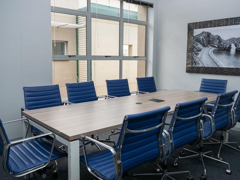 A professional office boardroom featuring a large central meeting table, blue ergonomic chairs, and large windows providing natural light.
