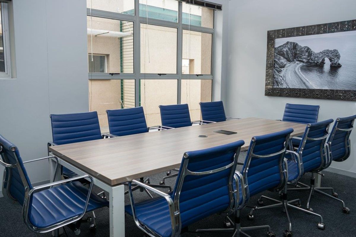 A professional office boardroom featuring a large central meeting table, blue ergonomic chairs, and large windows providing natural light.