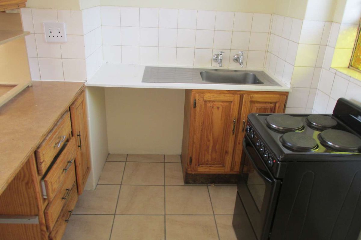 A functional kitchen area featuring wooden cabinetry, tiled splashbacks, a stainless steel sink, and tiled flooring, offering potential for modernization.