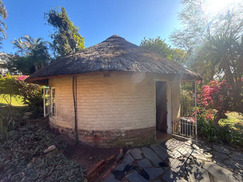 A detached circular cottage featuring a traditional thatched roof, brick construction, and a stone-paved entrance area set within a garden.