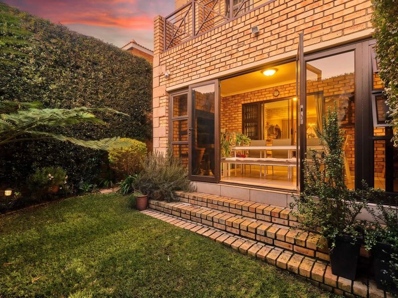 Rear view of a face brick townhouse featuring a landscaped garden, brick steps, and double doors leading into the living area with an upper balcony.