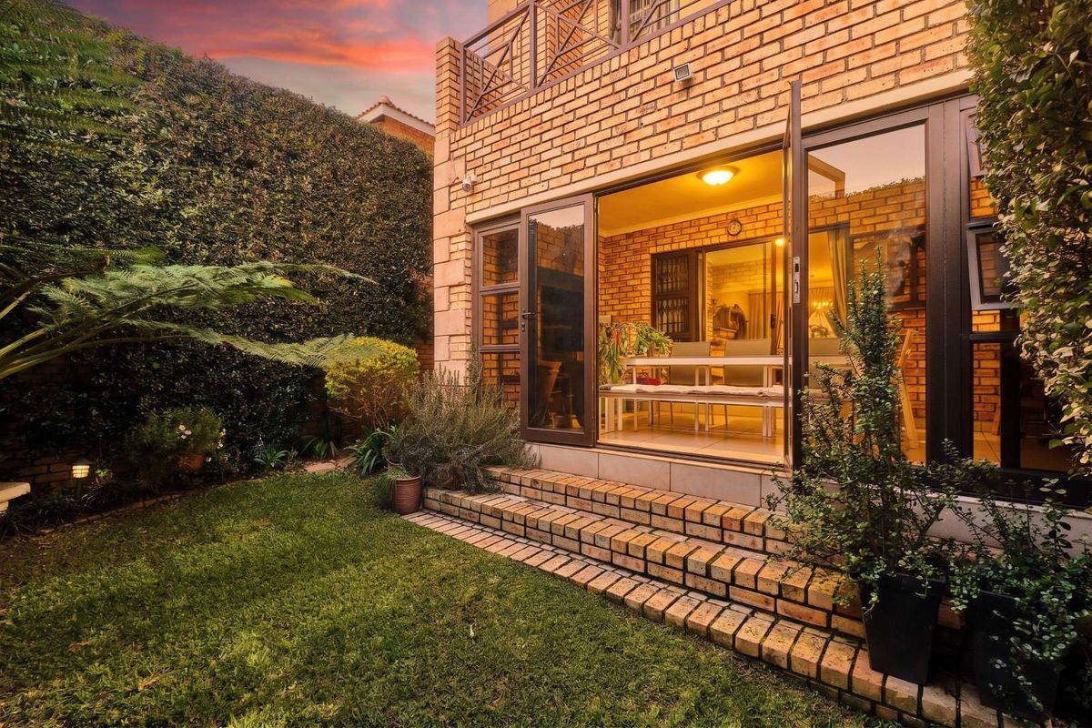 Rear view of a face brick townhouse featuring a landscaped garden, brick steps, and double doors leading into the living area with an upper balcony.