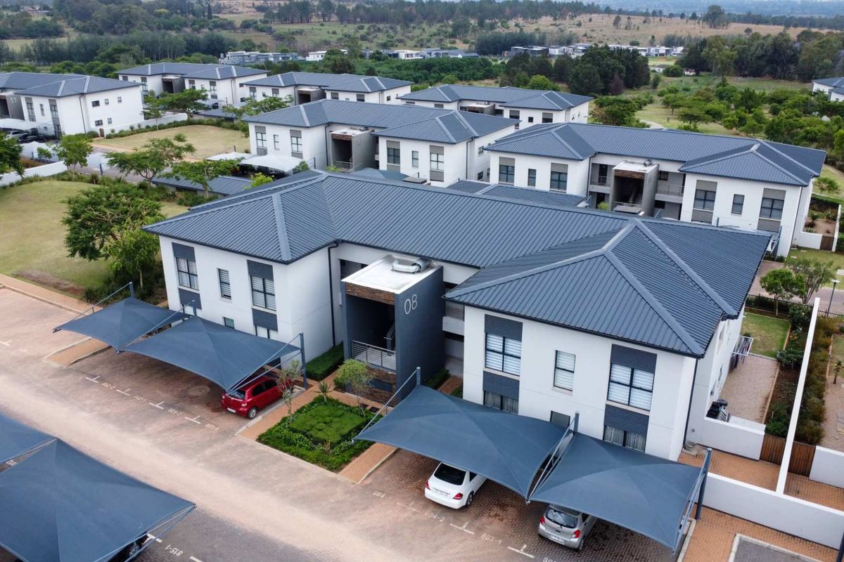 Aerial view of a modern apartment complex featuring pitched metal roofs, paved parking areas with shade cloth carports, and landscaped communal grounds.