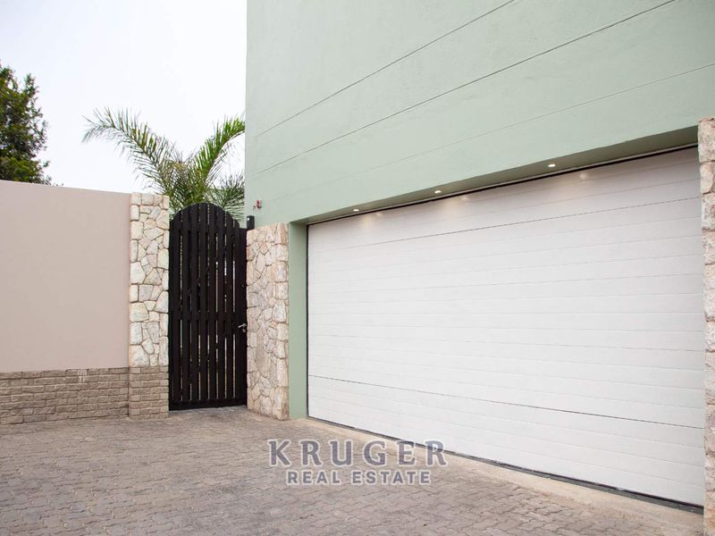 Modern garage exterior featuring a white sectional door, stone cladding accents, and integrated recessed lighting, complemented by a paved driveway and pedestrian gate.