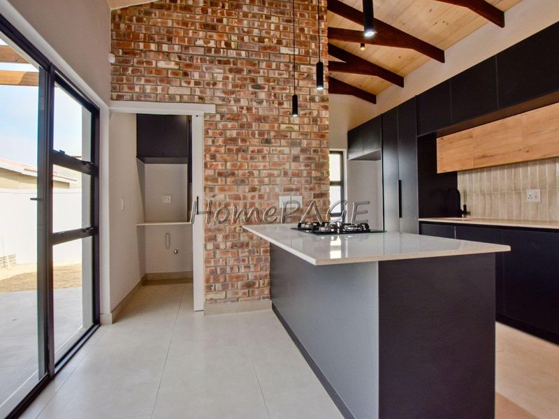 Modern kitchen featuring an exposed brick feature wall, timber beamed ceiling, sleek dark cabinetry, stone island countertop, and integrated gas hob.