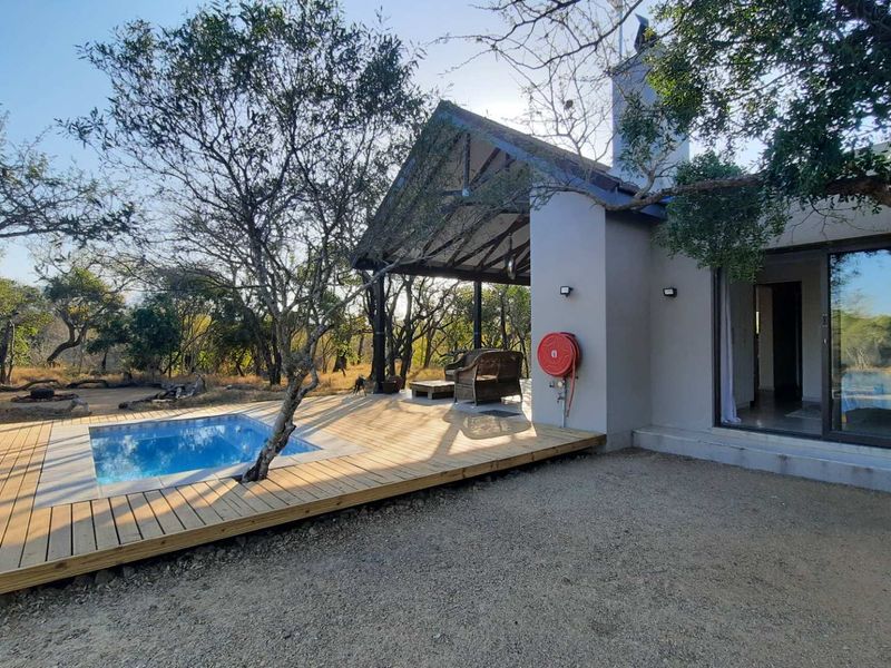 Outdoor entertainment area featuring a swimming pool, timber decking, and a covered patio with a fireplace, set within a natural bushveld environment.