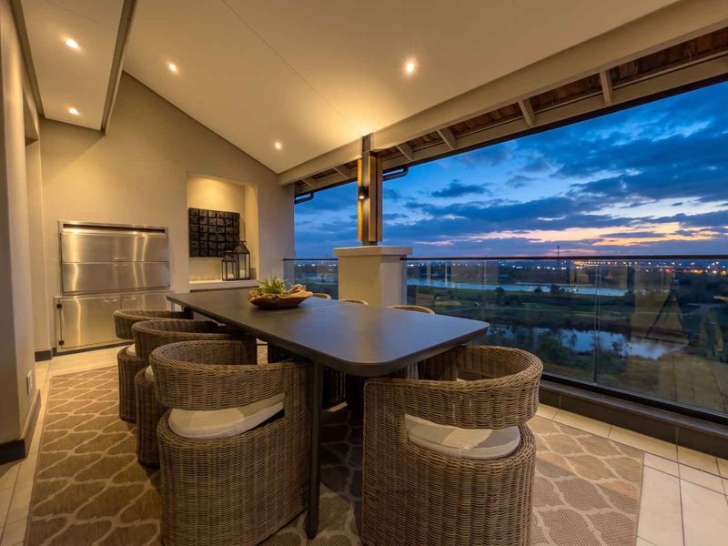 A modern covered balcony featuring a built-in braai, glass balustrade, and scenic sunset views over the surrounding landscape.