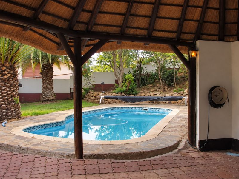 Outdoor pool area featuring a thatched lapa, stone paving, and a well-maintained swimming pool set within a landscaped garden.
