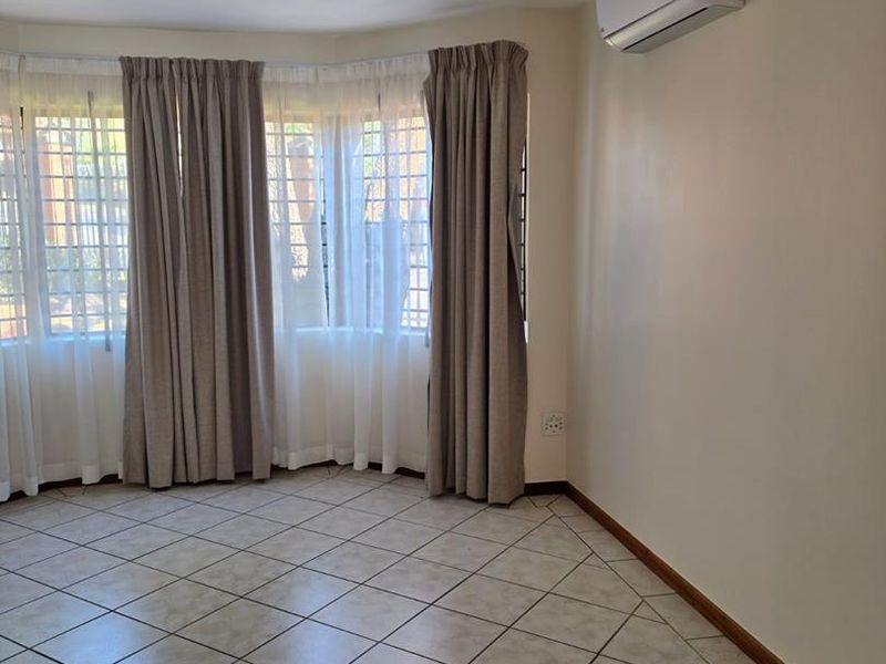 A bright, empty living room featuring neutral ceramic floor tiles, a wall-mounted air conditioning unit, and large windows fitted with security bars.