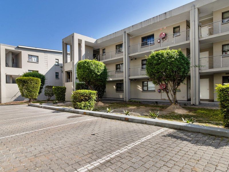 Exterior view of a multi-story apartment complex featuring paved parking bays, landscaped garden areas, and balconies with metal railings.