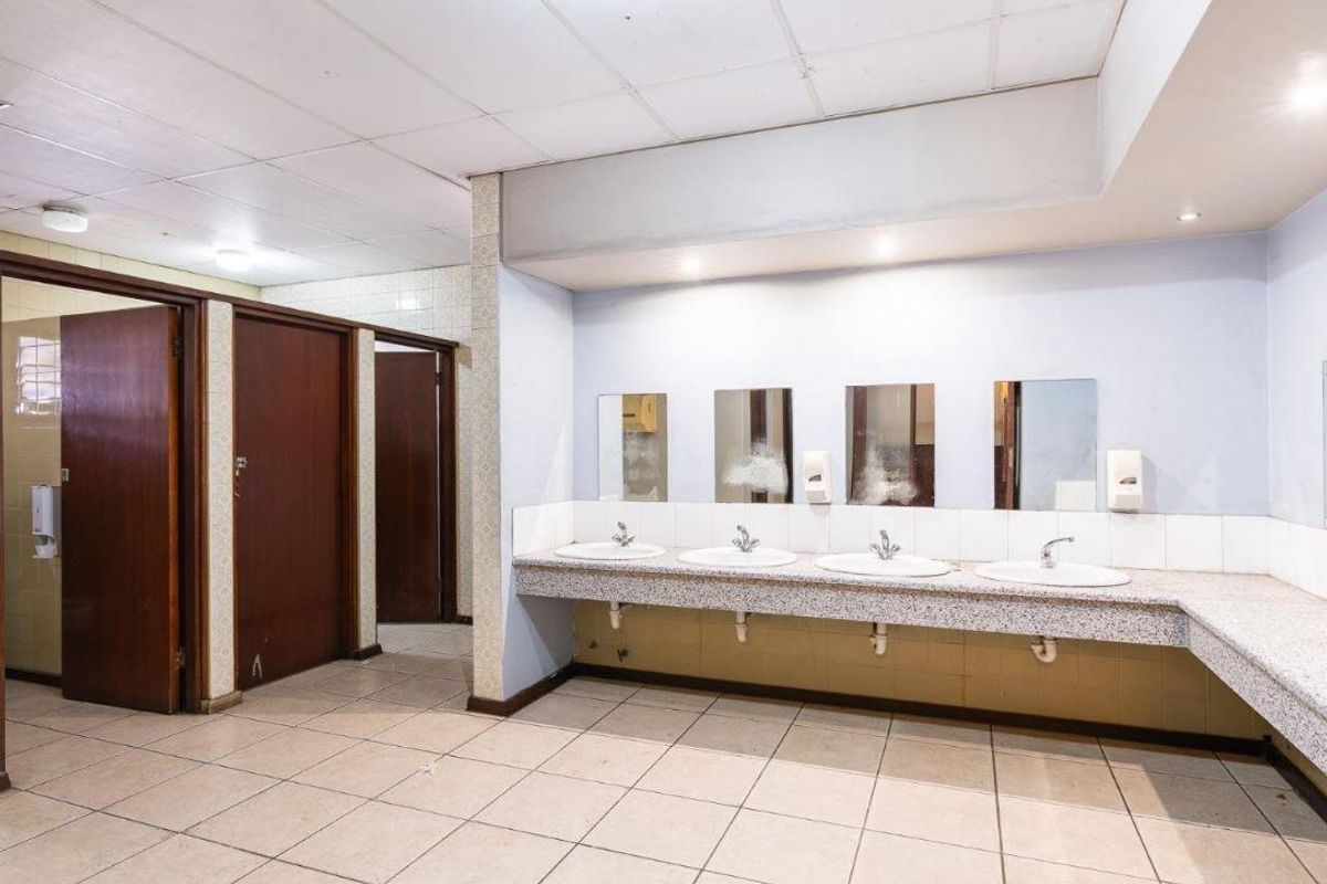 A commercial-style communal bathroom featuring a long vanity with multiple integrated sinks, tiled flooring, and several private toilet cubicles.