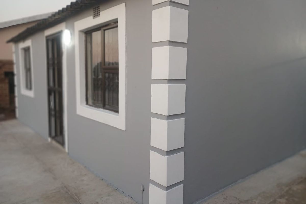 A side view of the house exterior featuring grey painted walls with white corner quoins, aluminium window frames, and a security gate.